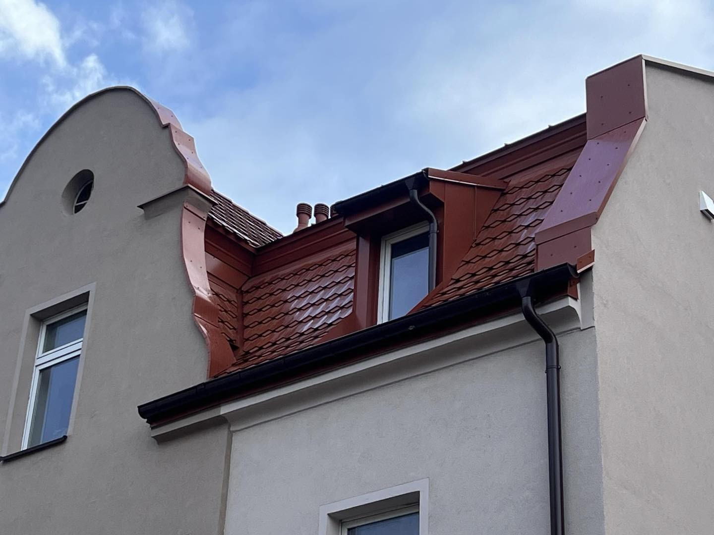 Czerwony dach z blachy na budynku z szarą elewacją, widoczne okna dachowe i rynny. Detal architektoniczny.