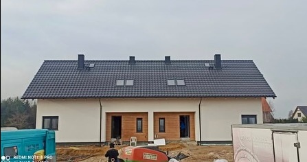 Nowoczesny dom z ciemnoszarą dachówką, widoczne okna dachowe i kominy. Jasna elewacja, częściowo wykończone otoczenie budynku. Ujęcie z poziomu terenu.