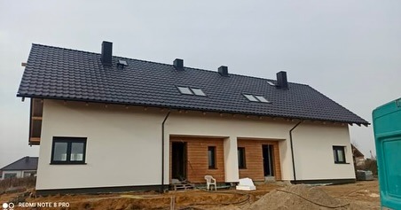 Nowoczesny dom z ciemną, ukończoną dachówką i oknami dachowymi. Widoczne kominy i drewniane elementy elewacji. Teren wokół w trakcie prac wykończeniowych.