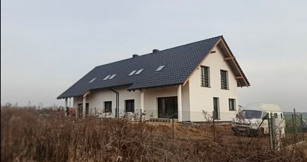 Nowoczesny dom jednorodzinny z ciemnoszarym dachem z dachówki ceramicznej, widok z poziomu gruntu, na pierwszym planie sucha trawa, biały bus zaparkowany obok.