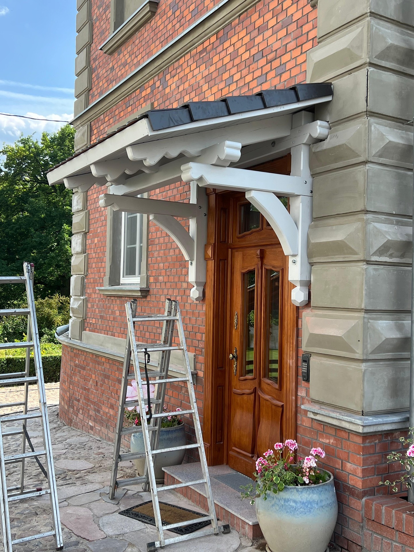 Wykończone zadaszenie nad wejściem do domu z cegły, wsparte na białych, zdobionych wspornikach. Dwie drabiny opierają się o elewację, a w donicach kwitną kwiaty.