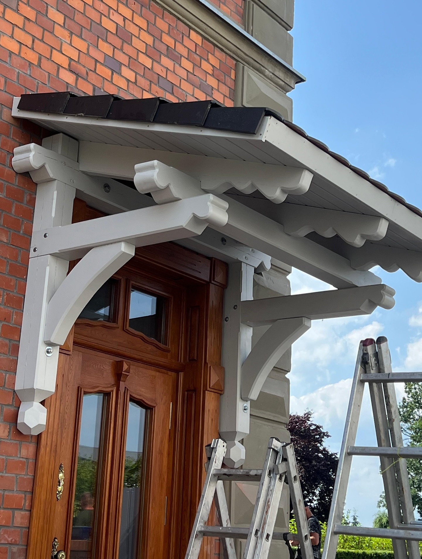 Zadaszenie nad wejściem z brązową dachówką i dekoracyjnymi, białymi elementami drewnianymi. Widoczna drabina oparta o ścianę z cegły.