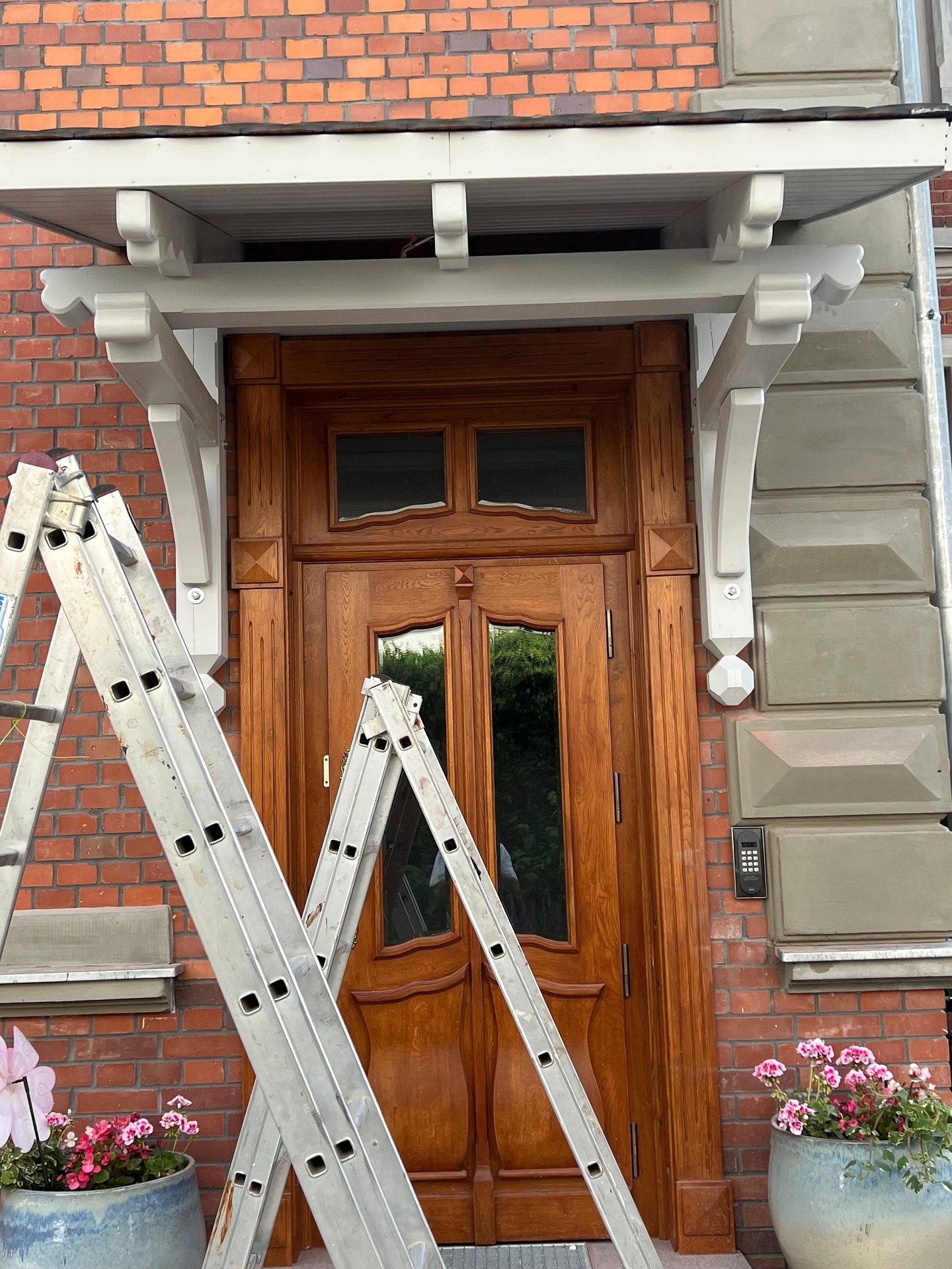Drewniane drzwi wejściowe z daszkiem, widziane z dołu, z rozstawionymi drabinami i kwiatami w donicach po bokach. Czerwona cegła w tle.