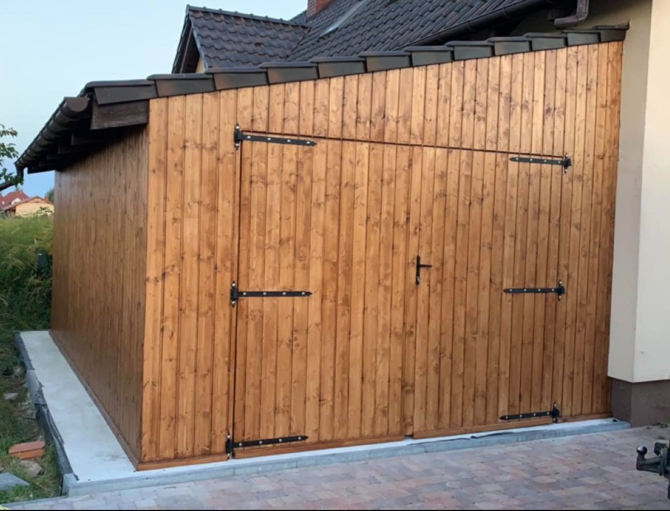 Drewniana konstrukcja z dachem krytym brązową dachówką, prawdopodobnie garaż lub budynek gospodarczy, z czarnymi okuciami, na tle domu jednorodzinnego.