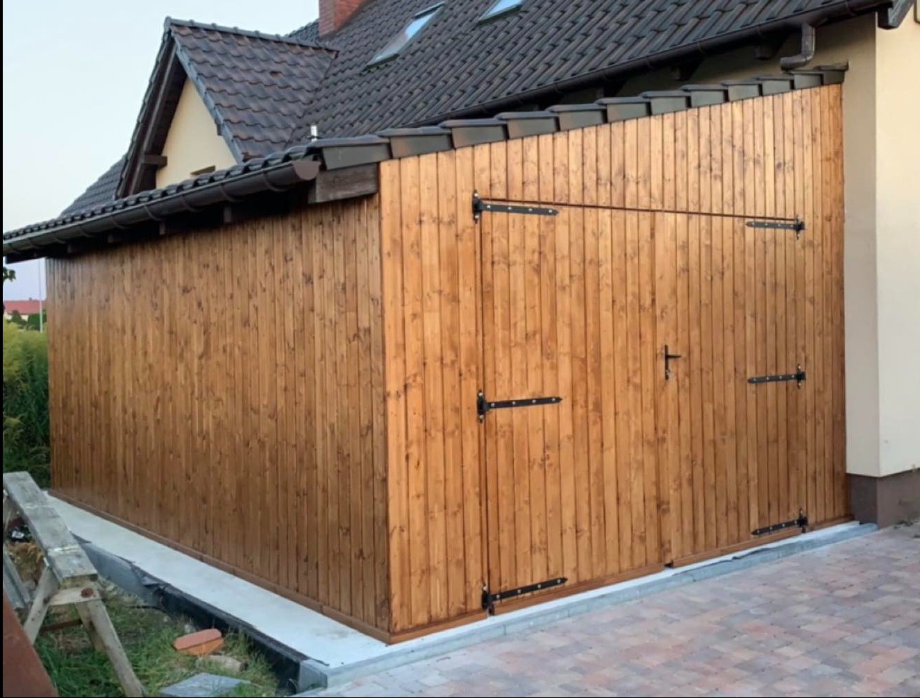 Drewniana dobudówka przylegająca do budynku z dachem krytym dachówką. Widoczne czarne okucia i rynny. Na pierwszym planie fragment drabiny.