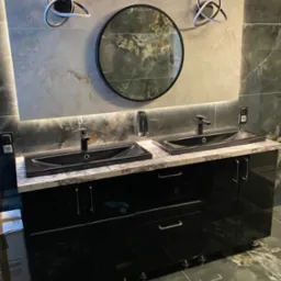 Nowoczesna łazienka z czarnymi szafkami pod umywalki, okrągłym lustrem i dekoracyjnym oświetleniem LED na ścianie z płytek imitujących marmur.