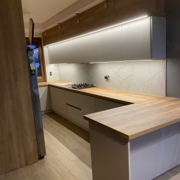 PIOTR BARTKIEWICZ MEBLE - Nowoczesna kuchnia z drewnianym blatem i białymi szafkami podświetlonymi ledami. Widoczny fragment lodówki i płyta grzewcza. Parkiet na podłodze.
