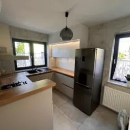 Jasna kuchnia z drewnianymi blatami i marmurowymi płytkami. Nowoczesny design, czarna lodówka i okno z firanką. Oświetlenie punktowe i wisząca lampa.