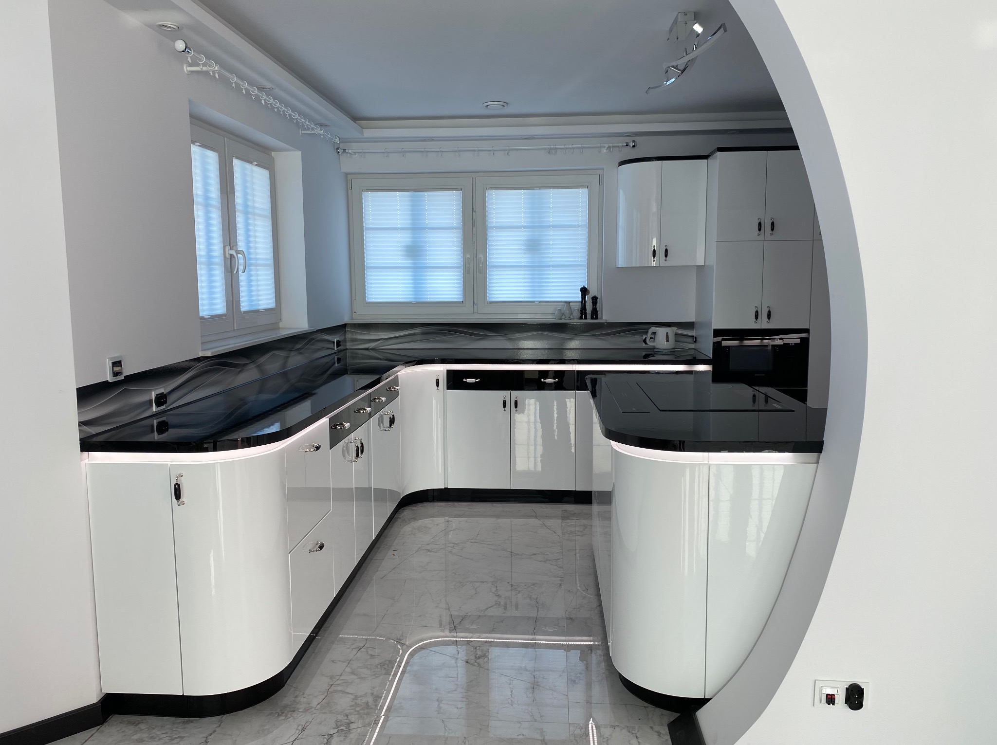 Elegancka, nowoczesna kuchnia z białymi, błyszczącymi szafkami i czarnym blatem. Marmurowa podłoga i designerskie oświetlenie tworzą luksusowy wygląd.