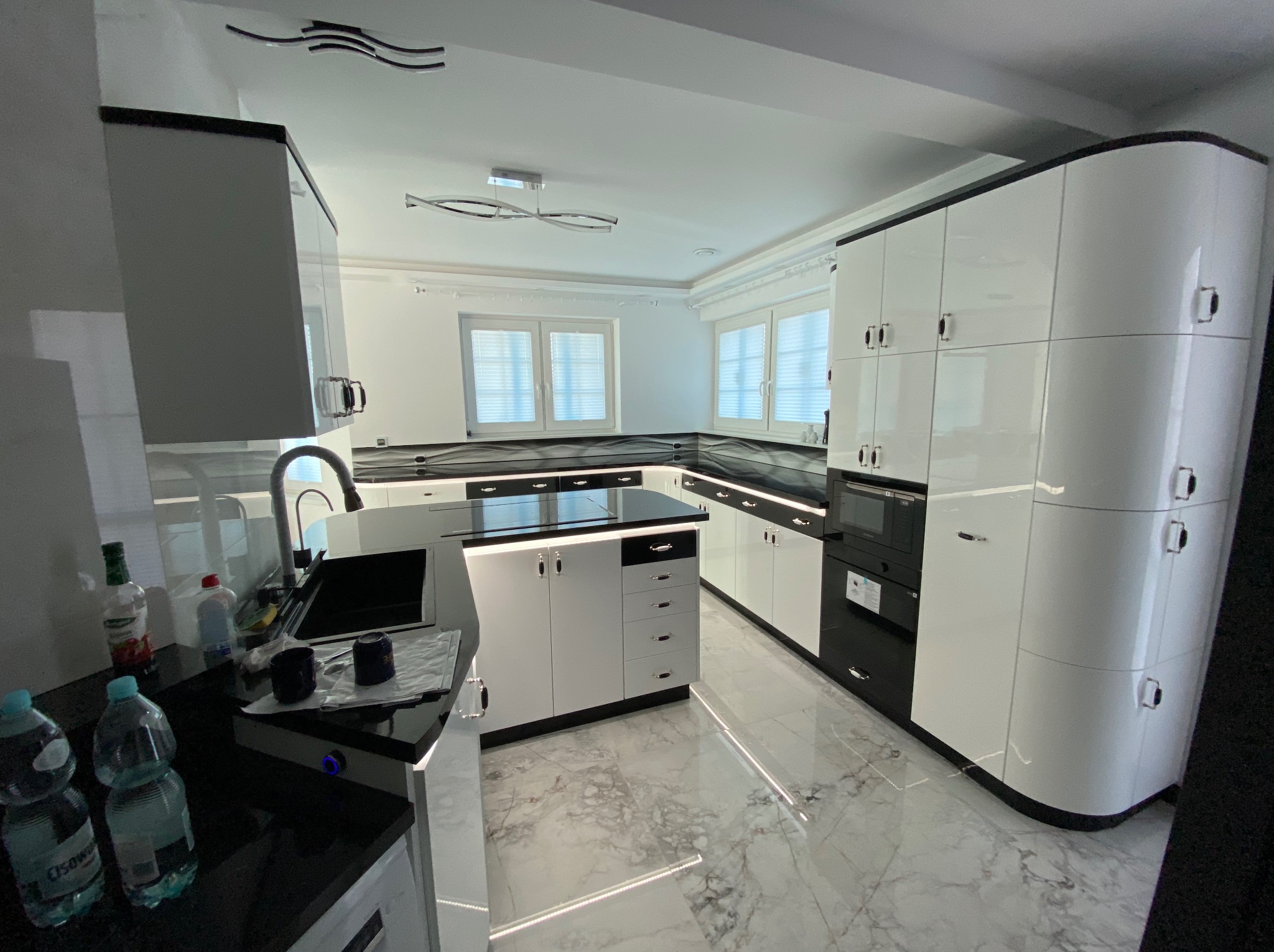 Biała, nowoczesna kuchnia z czarnymi akcentami i marmurową podłogą. Widoczne szafki z połyskiem, wyspa kuchenna i okno z żaluzjami. Całość tworzy elegancki i funkcjonalny design.