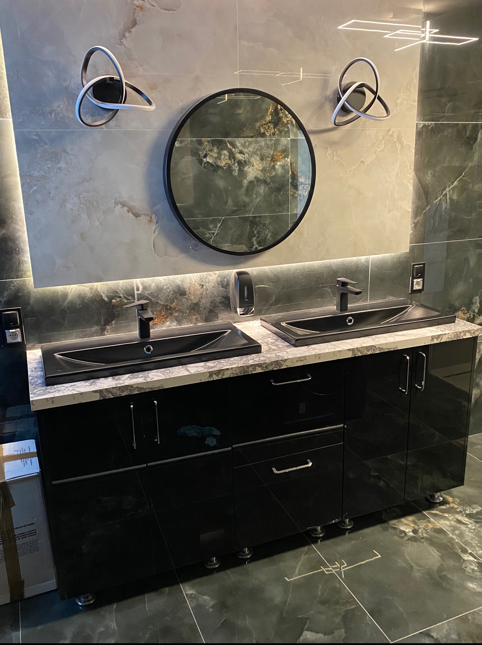 Nowoczesna łazienka z czarnymi szafkami pod umywalki, okrągłym lustrem i dekoracyjnym oświetleniem LED na ścianie z płytek imitujących marmur.
