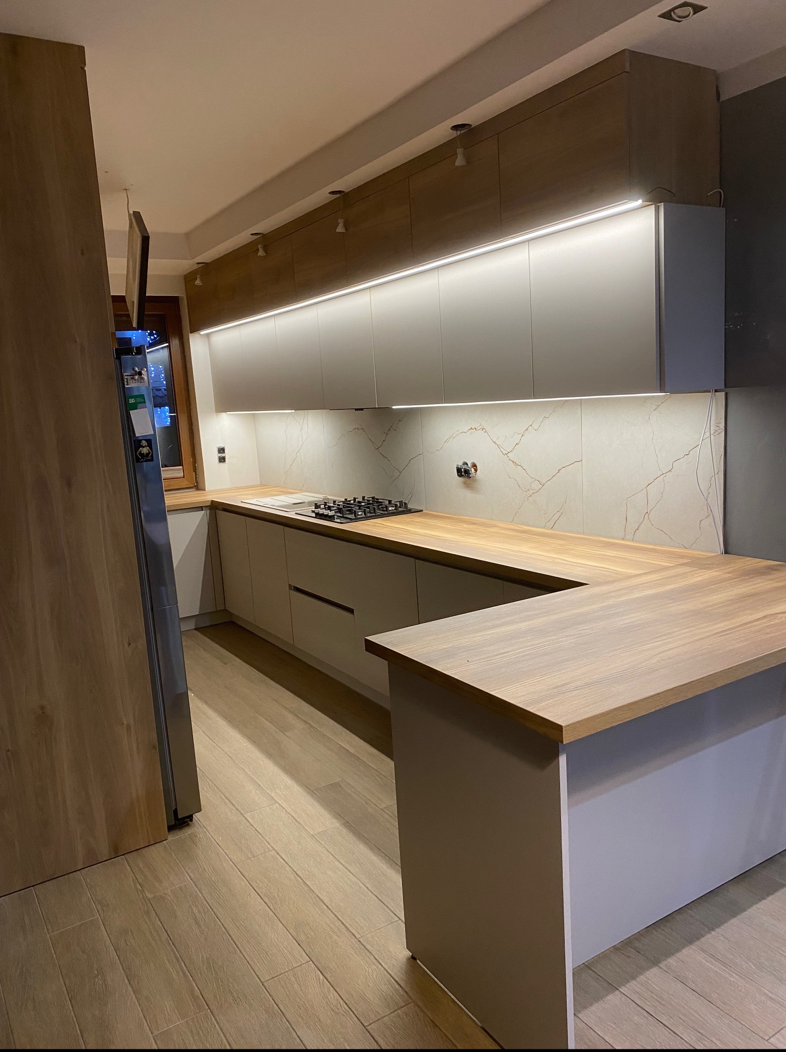 Nowoczesna kuchnia z drewnianym blatem i białymi szafkami podświetlonymi ledami. Widoczny fragment lodówki i płyta grzewcza. Parkiet na podłodze.
