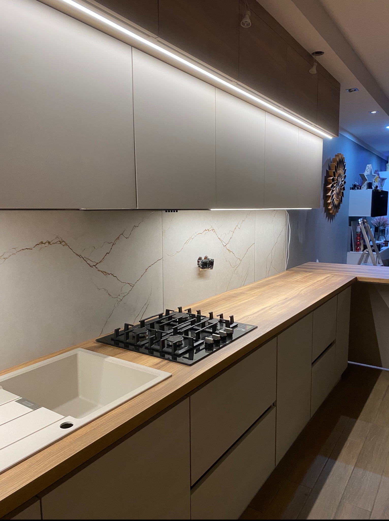 Nowoczesna kuchnia z drewnianym blatem, jasnymi szafkami i podświetleniem LED. Widoczna płyta gazowa i zlewozmywak. Marmurowe wykończenie ściany nad blatem.