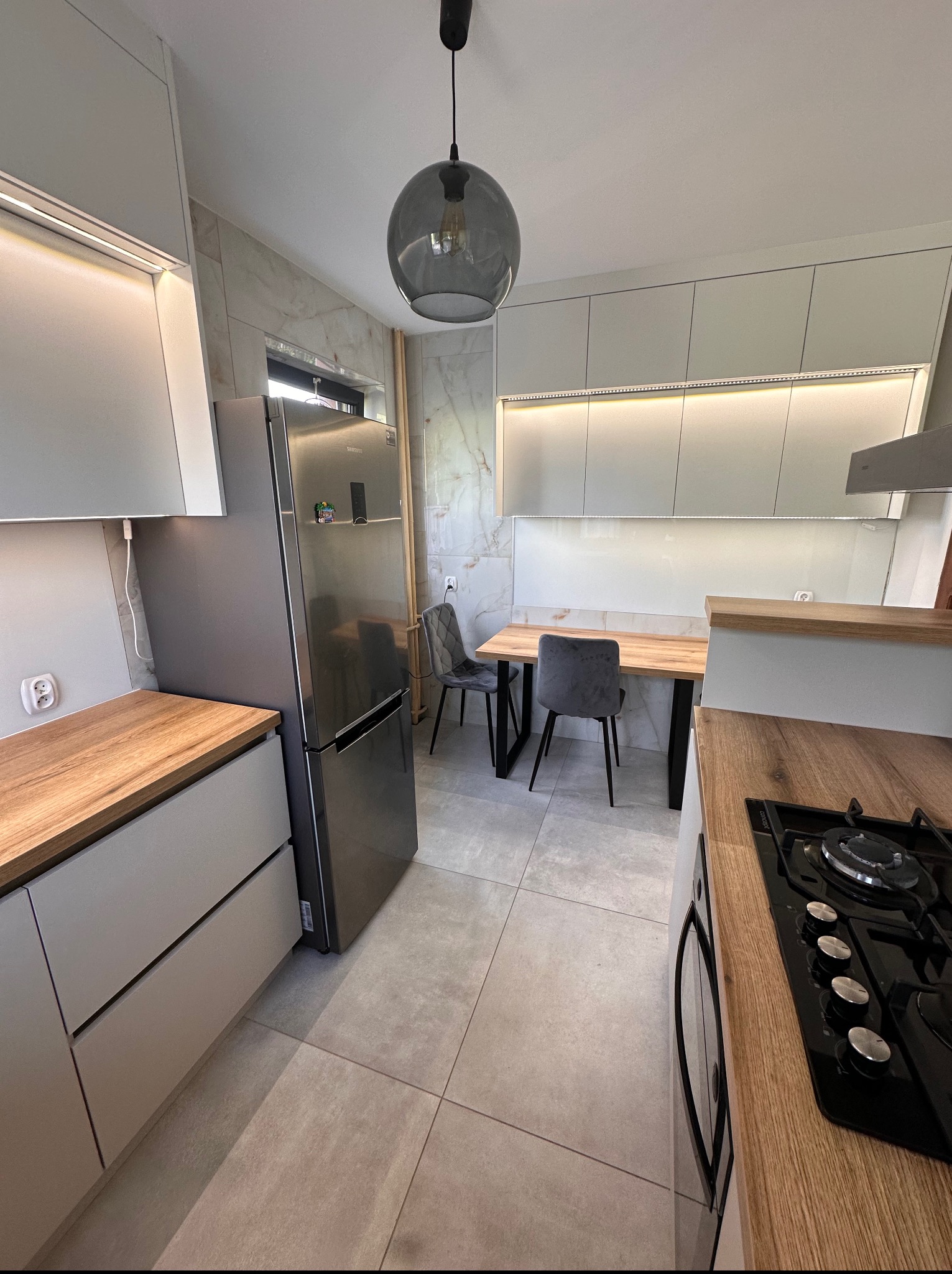 Jasna kuchnia z drewnianym blatem, szarymi szafkami i nowoczesnym oświetleniem LED. Na pierwszym planie widoczna płyta gazowa. W tle stół z krzesłami.