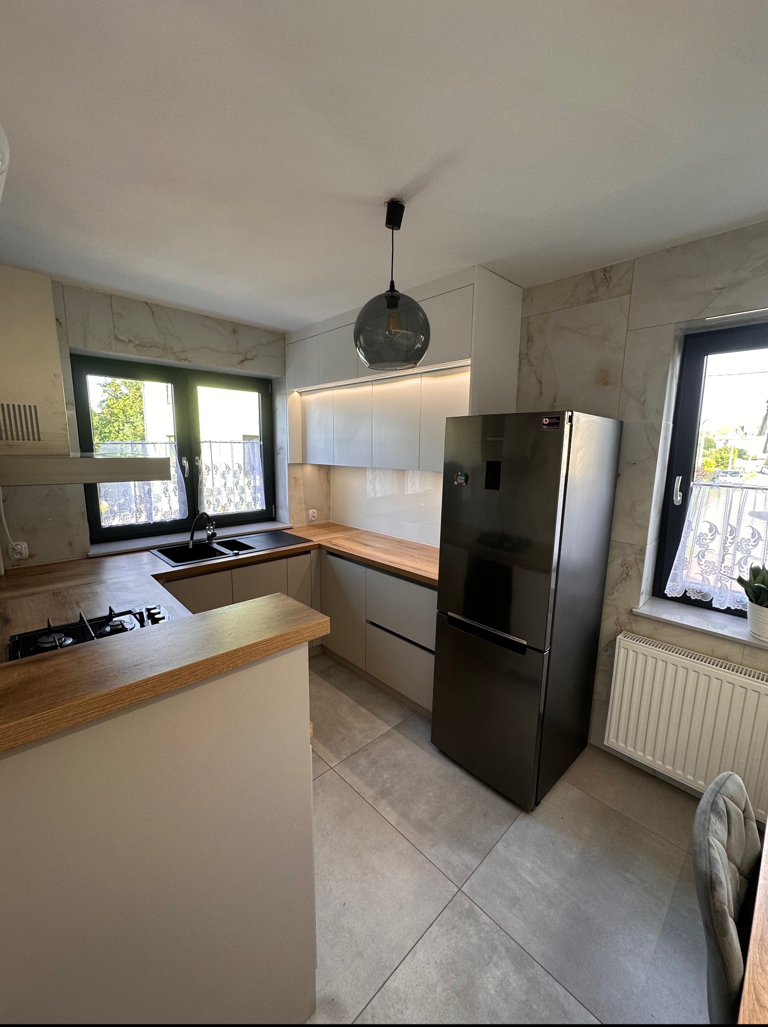 Jasna kuchnia z drewnianymi blatami i marmurowymi płytkami. Nowoczesny design, czarna lodówka i okno z firanką. Oświetlenie punktowe i wisząca lampa.