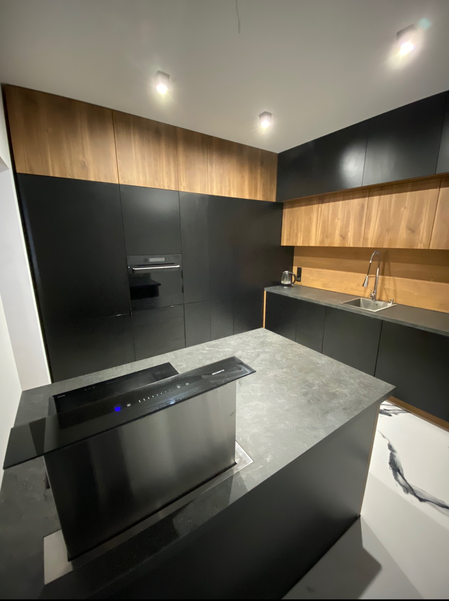 Nowoczesna kuchnia z czarnymi szafkami i drewnianymi akcentami, wyspa kuchenna z płytą indukcyjną i pochłaniaczem. Minimalistyczny design, oświetlenie punktowe.