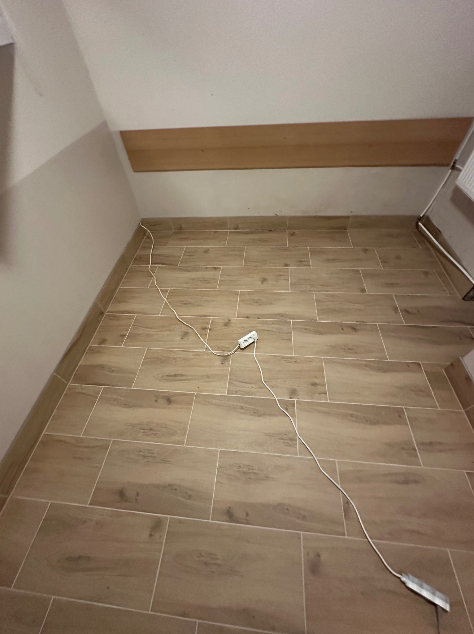 Ukończona podłoga z płytek imitujących drewno w skosie poddasza. Widoczny kabel zasilający i listwa montażowa na ścianie. Ciepła kolorystyka.