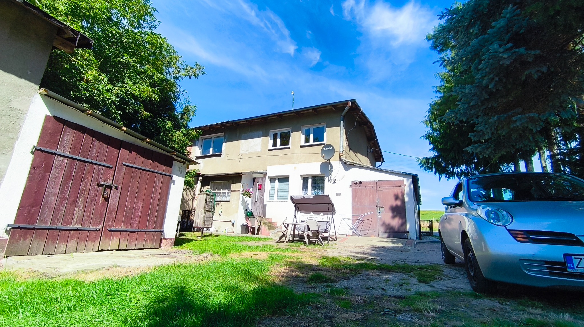 Dwukondygnacyjny dom z podwórkiem, garażem i samochodem w słoneczny dzień. Widoczne meble ogrodowe i antena satelitarna. Ujęcie z niskiej perspektywy.