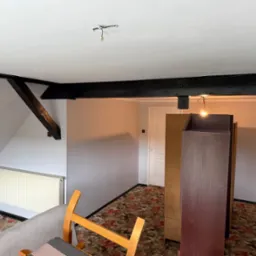 Poddasze po malowaniu: świeże, szare ściany, ciemne belki stropowe, grzejnik i fragmenty mebli. Widoczna instalacja elektryczna bez oprawy.