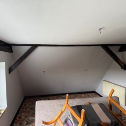 Mateusz Bury Krzysztof Czerny - Poddasze ze skośnym sufitem pomalowanym na biało, z widocznymi, ciemnymi belkami stropowymi. Na oknie napoje i naczynia. Widoczny fragment mebli i grzejnik.