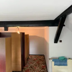 Wnętrze poddasza po odświeżeniu: pomalowane ściany na biało, widoczne drewniane, pomalowane na czarno belki stropowe, grzejnik z ręcznikami i meble w korytarzu.