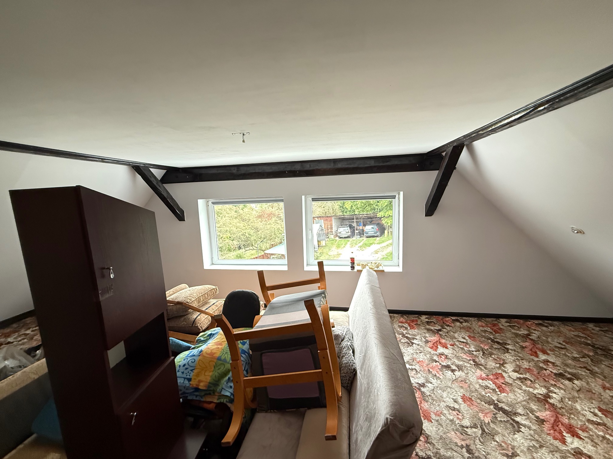 Poddasze przed remontem: białe ściany, drewniane belki, meble przesunięte na środek, dywan w liście. Widok z okna na zieleń i zaparkowane samochody.