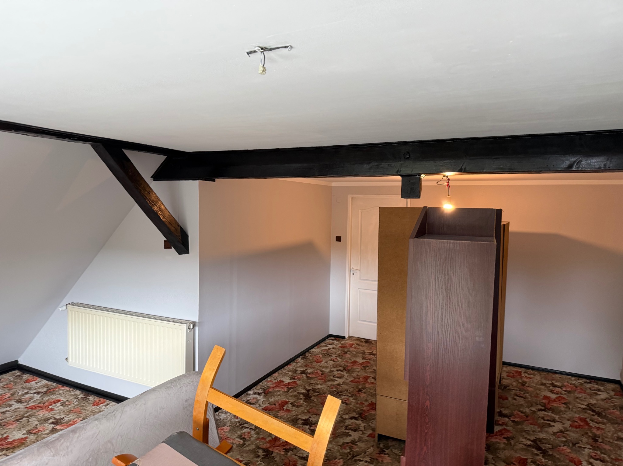 Poddasze po malowaniu: świeże, szare ściany, ciemne belki stropowe, grzejnik i fragmenty mebli. Widoczna instalacja elektryczna bez oprawy.