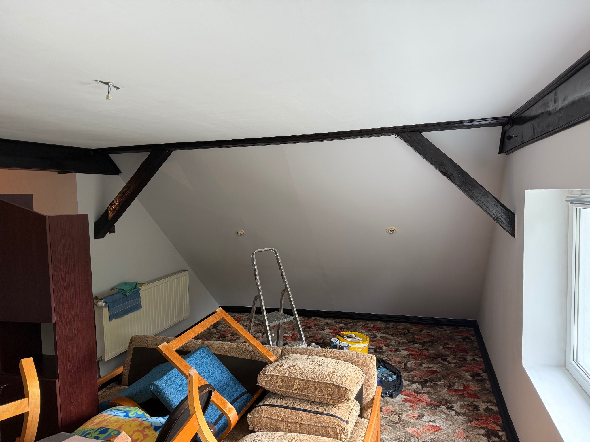 Poddasze po malowaniu z czarnymi belkami, widoczna drabina, meble w nieładzie. Odświeżone wnętrze w trakcie porządkowania.
