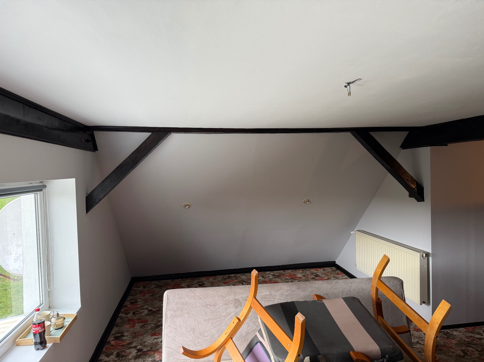 Poddasze ze skośnym sufitem pomalowanym na biało, z widocznymi, ciemnymi belkami stropowymi. Na oknie napoje i naczynia. Widoczny fragment mebli i grzejnik.
