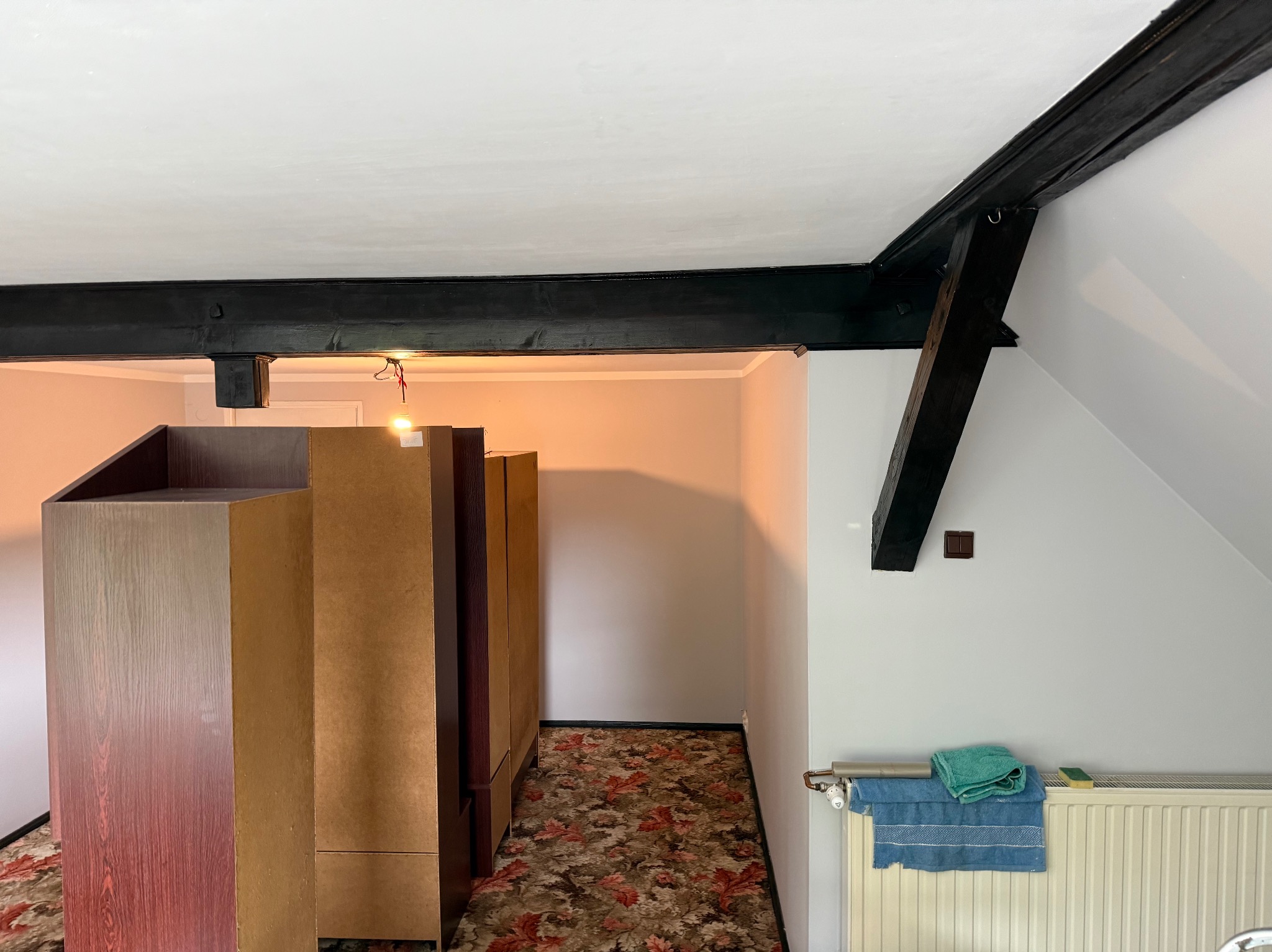 Wnętrze poddasza po odświeżeniu: pomalowane ściany na biało, widoczne drewniane, pomalowane na czarno belki stropowe, grzejnik z ręcznikami i meble w korytarzu.