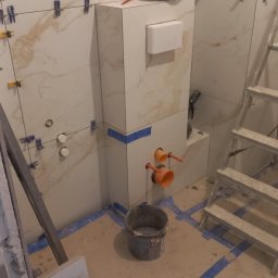 Montaż płytek imitujących marmur w łazience. Widoczne narzędzia, poziomice, krzyżyki dystansowe i fragment instalacji. Prace w toku, zabezpieczona podłoga.