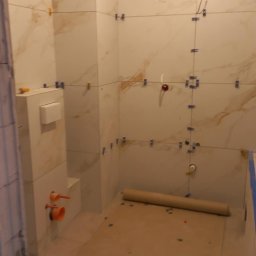 Łazienka w trakcie remontu: ułożone płytki imitujące marmur na ścianach, widoczne krzyżyki dystansowe, zabudowa WC, rury. Podłoga zabezpieczona kartonem.