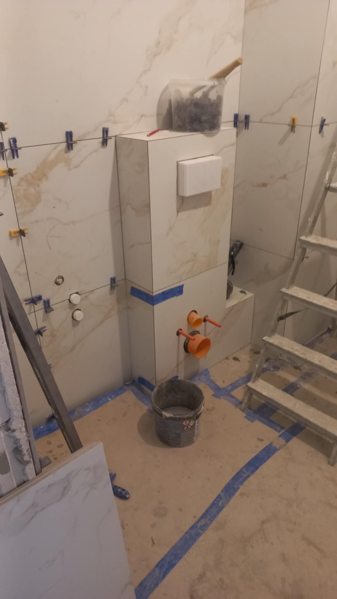 Montaż płytek imitujących marmur w łazience. Widoczne narzędzia, poziomice, krzyżyki dystansowe i fragment instalacji. Prace w toku, zabezpieczona podłoga.