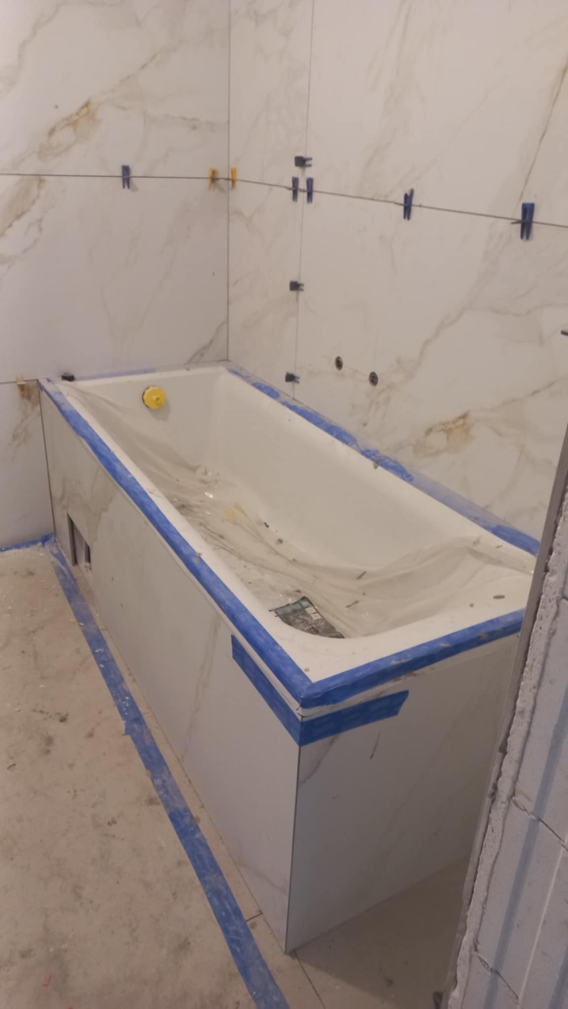 Obłożona marmurowymi płytkami wanna w trakcie prac wykończeniowych. Widoczne niebieskie taśmy zabezpieczające i krzyżyki glazurnicze na ścianach.
