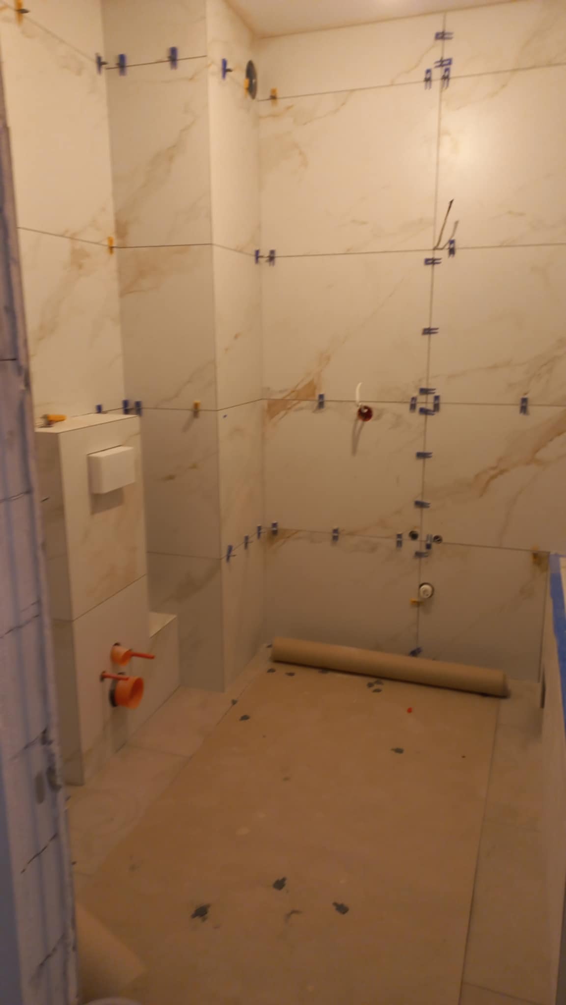 Łazienka w trakcie remontu: ułożone płytki imitujące marmur na ścianach, widoczne krzyżyki dystansowe, zabudowa WC, rury. Podłoga zabezpieczona kartonem.