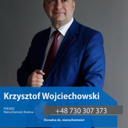 krzysztof Wojciechowski - Portret Krzysztofa Wojciechowskiego, doradcy ds. nieruchomości z Krakowa, w niebieskim garniturze i czerwonym krawacie, na szarym tle. Widoczny numer telefonu.