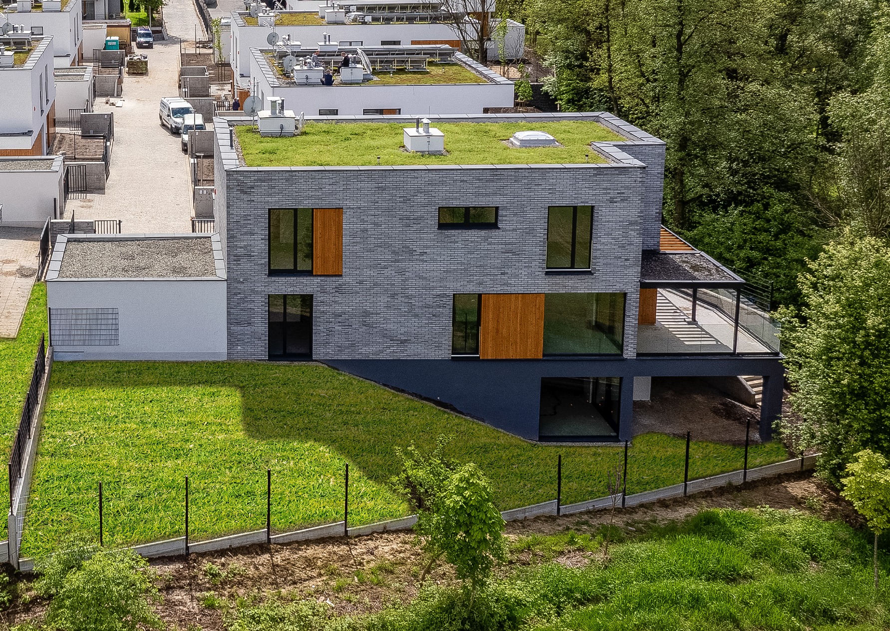 Nowoczesny dom z zielonym dachem i szarą elewacją z cegły, widok z góry. W tle inne budynki i drzewa. Zadbany trawnik otacza posesję.