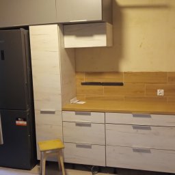 PPHU FARAM - Częściowo zamontowana kuchnia na wymiar z szarymi i drewnopodobnymi frontami, widoczny fragment sufitu podwieszanego i poziomica na górnej szafce.