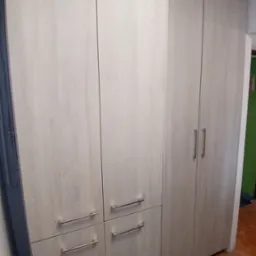 Jasna, drewnopodobna szafa z poziomymi uchwytami, umieszczona w rogu pokoju z widoczną podłogą i fragmentem ściany w tle. Fotografia wnętrza.