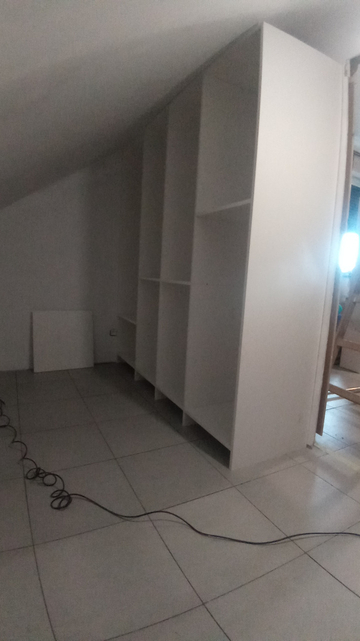 Biała szafa wnękowa z półkami, zamontowana w pokoju na poddaszu. Widoczny kabel na podłodze i fragment drabiny w tle.