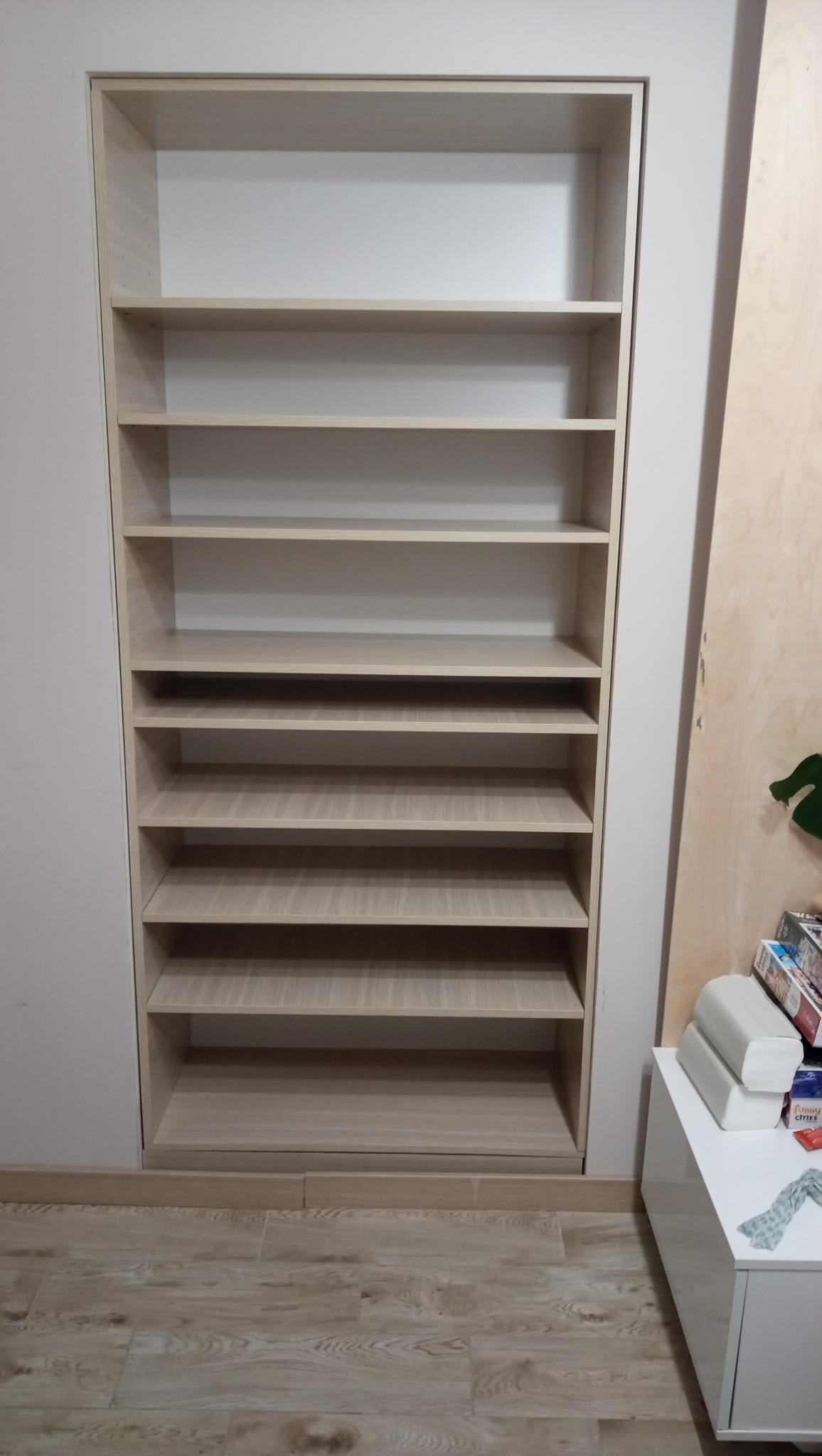 Jasna, drewniana szafa wnękowa z półkami, wbudowana w ścianę. Widoczne panele podłogowe i fragment białej komody z boku. Prosta, minimalistyczna forma.