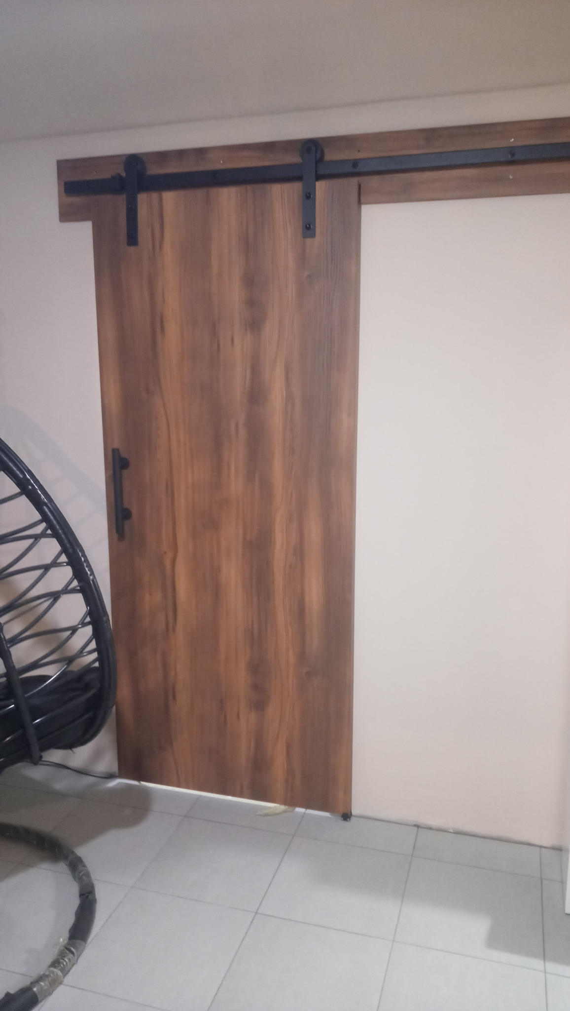 Drewniane drzwi przesuwne w stylu stodoły z czarnym okuciem, zamontowane na ścianie w jasnym pomieszczeniu. Widoczny fragment wiszącego fotela.