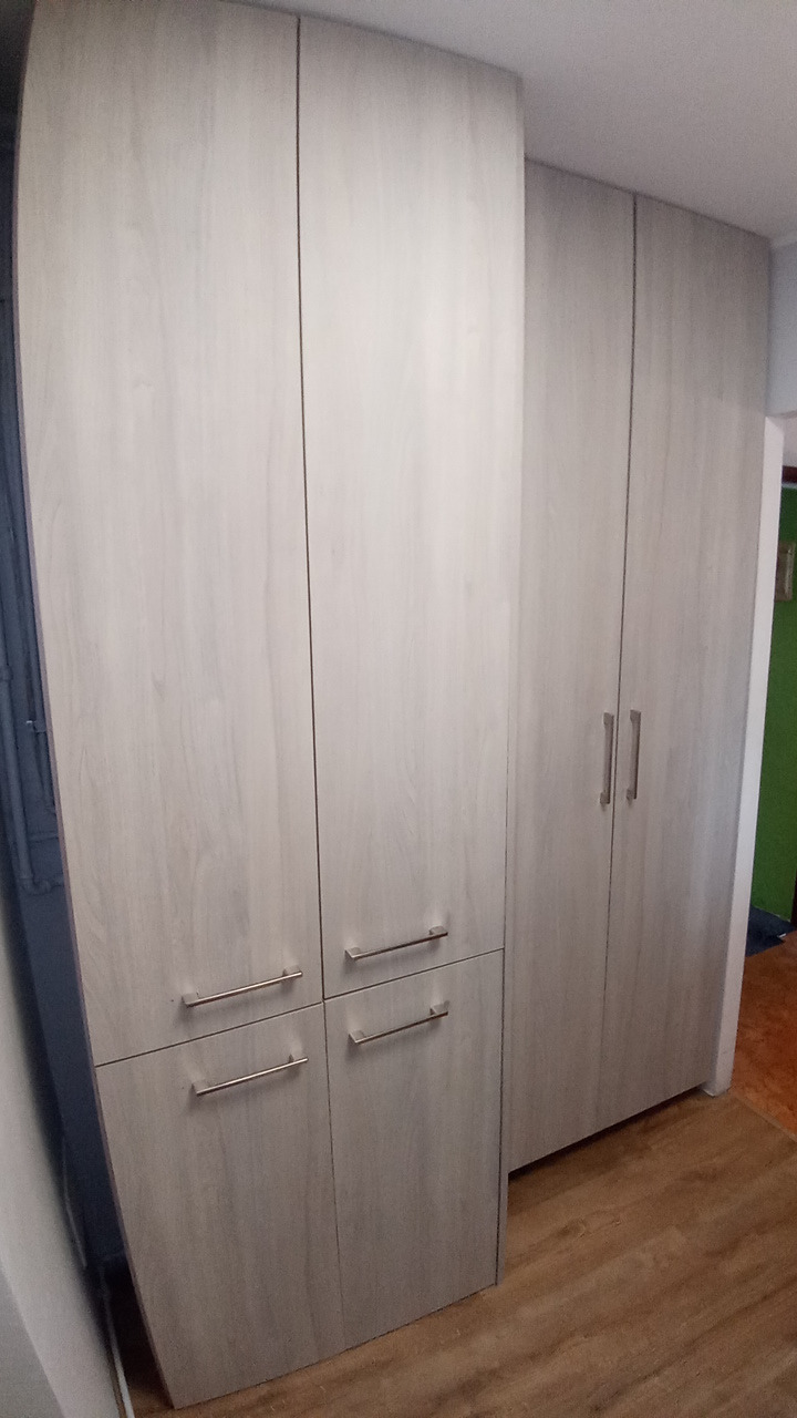 Jasna, drewnopodobna szafa z poziomymi uchwytami, umieszczona w rogu pokoju z widoczną podłogą i fragmentem ściany w tle. Fotografia wnętrza.