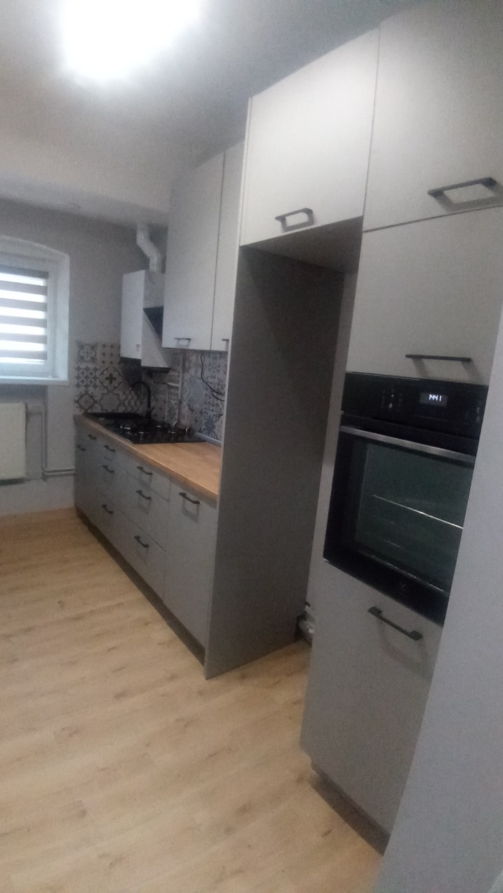 Szara kuchnia z drewnianym blatem i wzorzystymi płytkami nad kuchenką. Widoczny piekarnik w zabudowie i okno z żaluzjami.