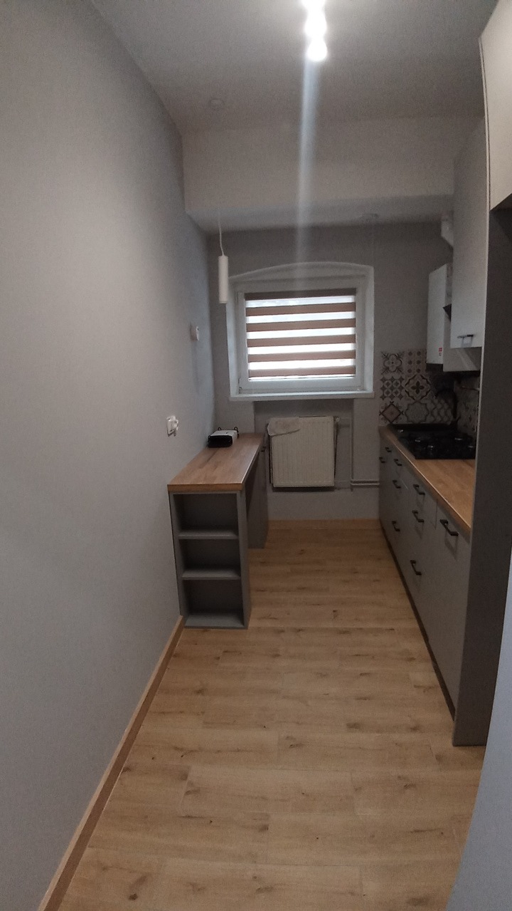 Szara kuchnia z drewnianym blatem i podłogą. Widok na okno z żaluzjami i zabudowę kuchenną z szufladami i piekarnikiem. Minimalistyczny design, wąskie pomieszczenie.