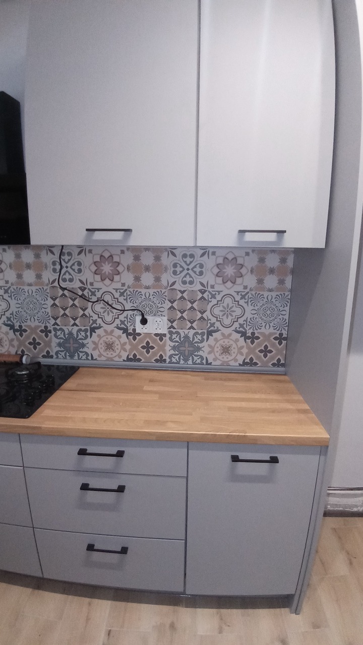 Szare szafki kuchenne z drewnianym blatem i dekoracyjną ścianą nad blatem. Widoczne czarne uchwyty i gniazdko elektryczne. Ujęcie z perspektywy lekko z dołu.