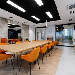 DDWNĘTRZA - Nowoczesna sala konferencyjna z długim stołem, pomarańczowymi krzesłami i dekoracyjną ścianą z efektem rdzy. Widoczne oświetlenie sufitowe i elementy dekoracyjne.