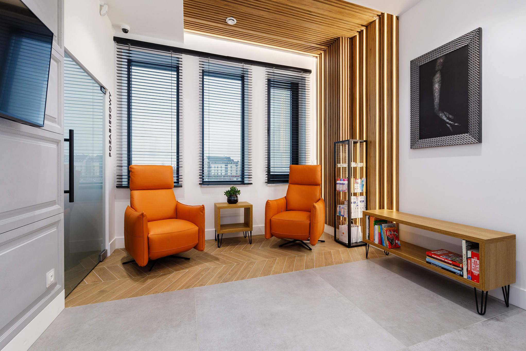 Stylowe wnętrze poczekalni z dwoma pomarańczowymi fotelami, drewnianym sufitem i ścianą z lamelami. Widoczne okna z żaluzjami i regał z książkami.
