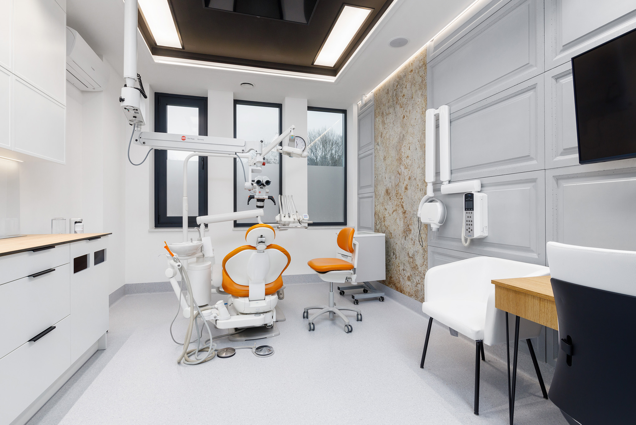 Nowoczesny gabinet dentystyczny z pomarańczowym fotelem, białymi meblami i panelami ściennymi. Jasne oświetlenie sufitowe i okna w tle. Minimalistyczny design.