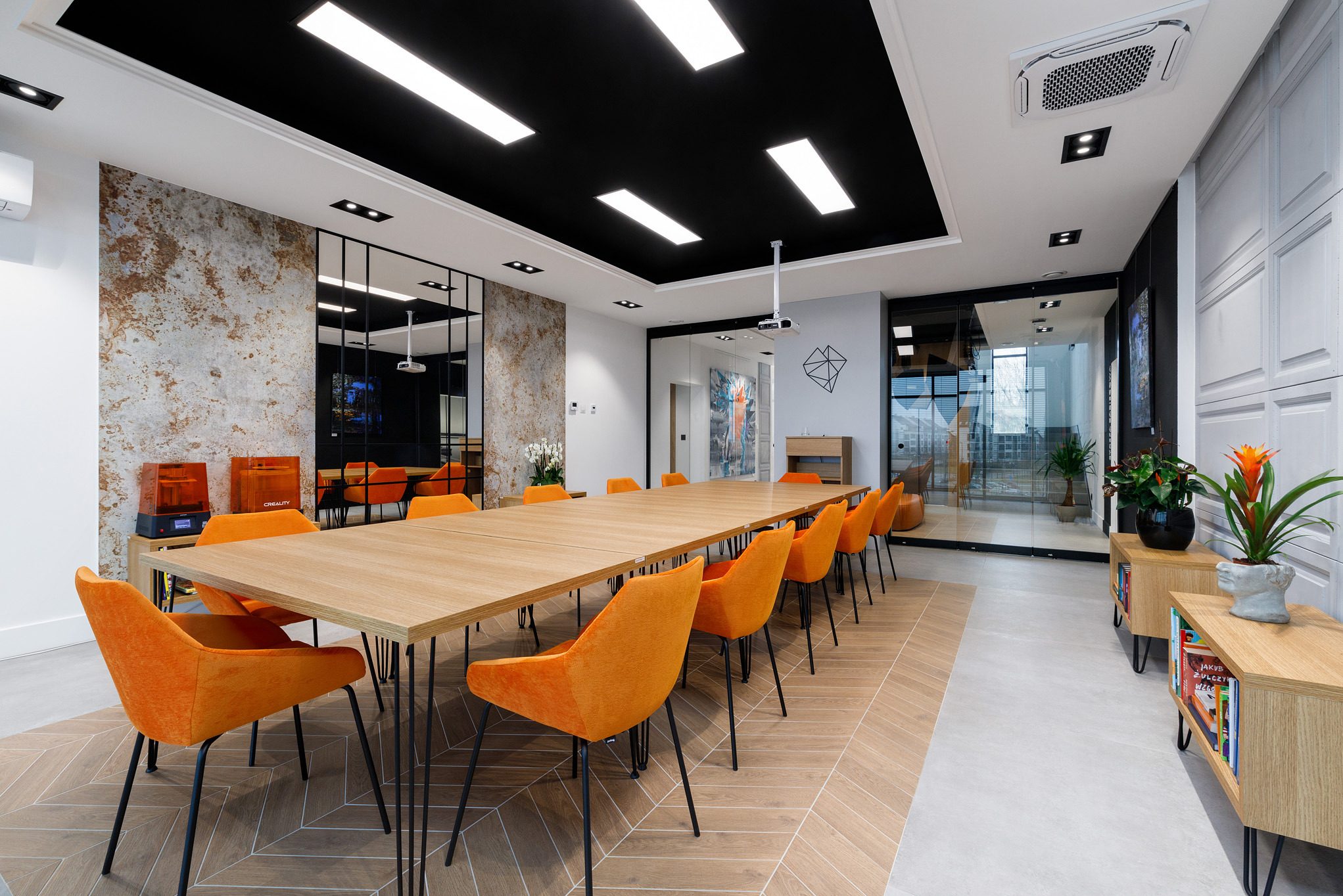 Nowoczesna sala konferencyjna z długim stołem, pomarańczowymi krzesłami i dekoracyjną ścianą z efektem rdzy. Widoczne oświetlenie sufitowe i elementy dekoracyjne.